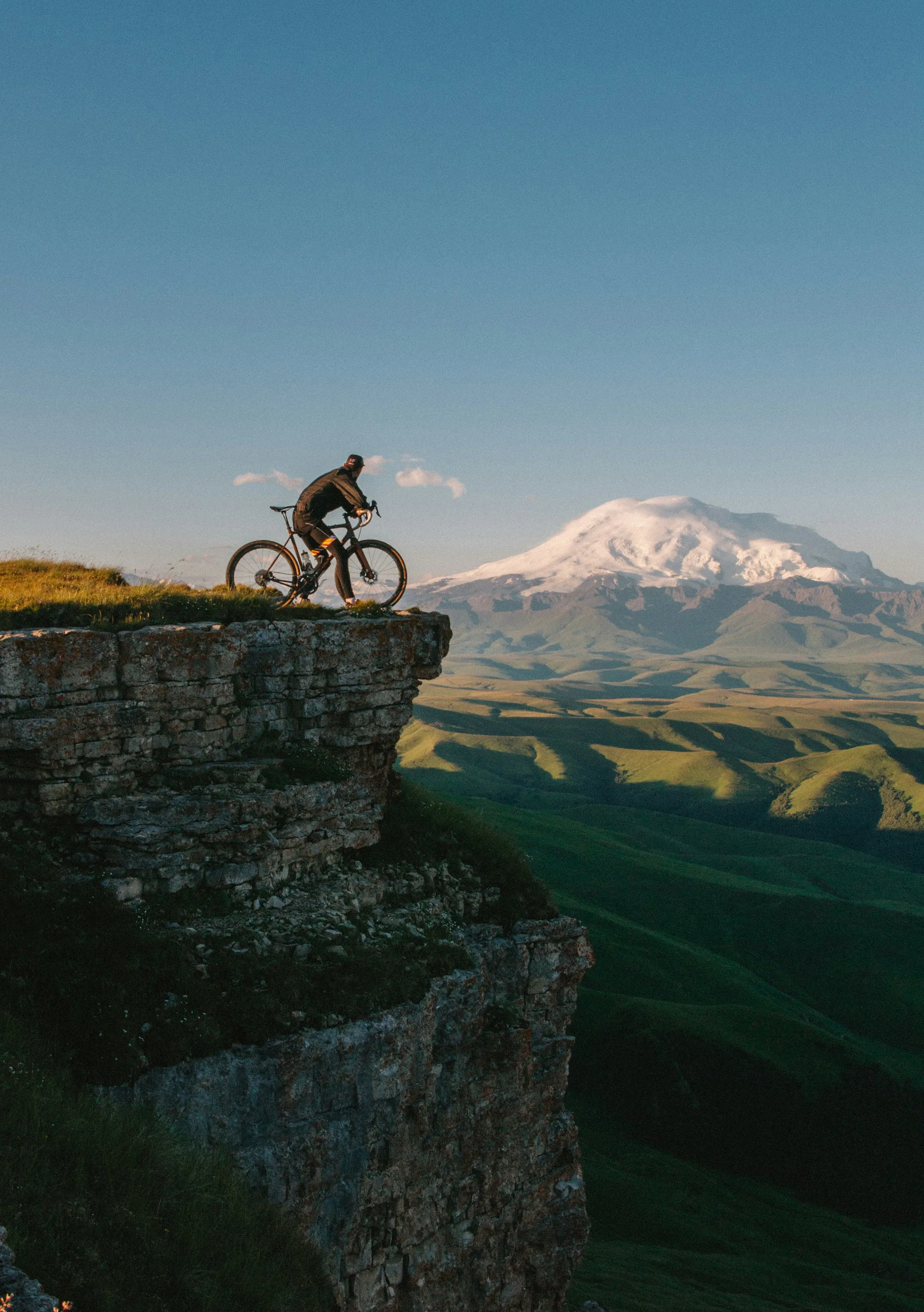 A cyclist riding along a cliff edge with a snowy volcano behind