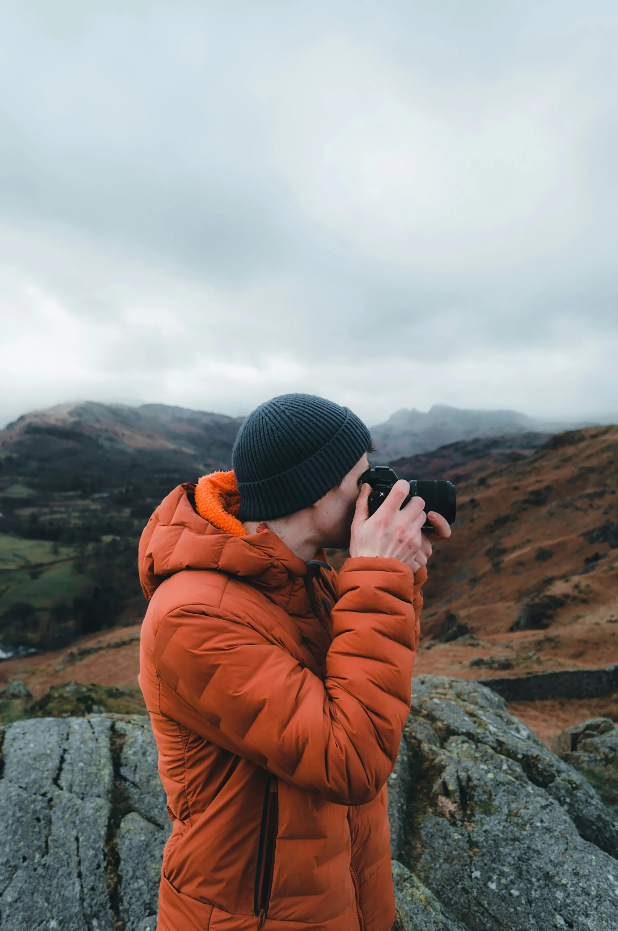 A man in an orange insulated jacket taking a photo in the hills