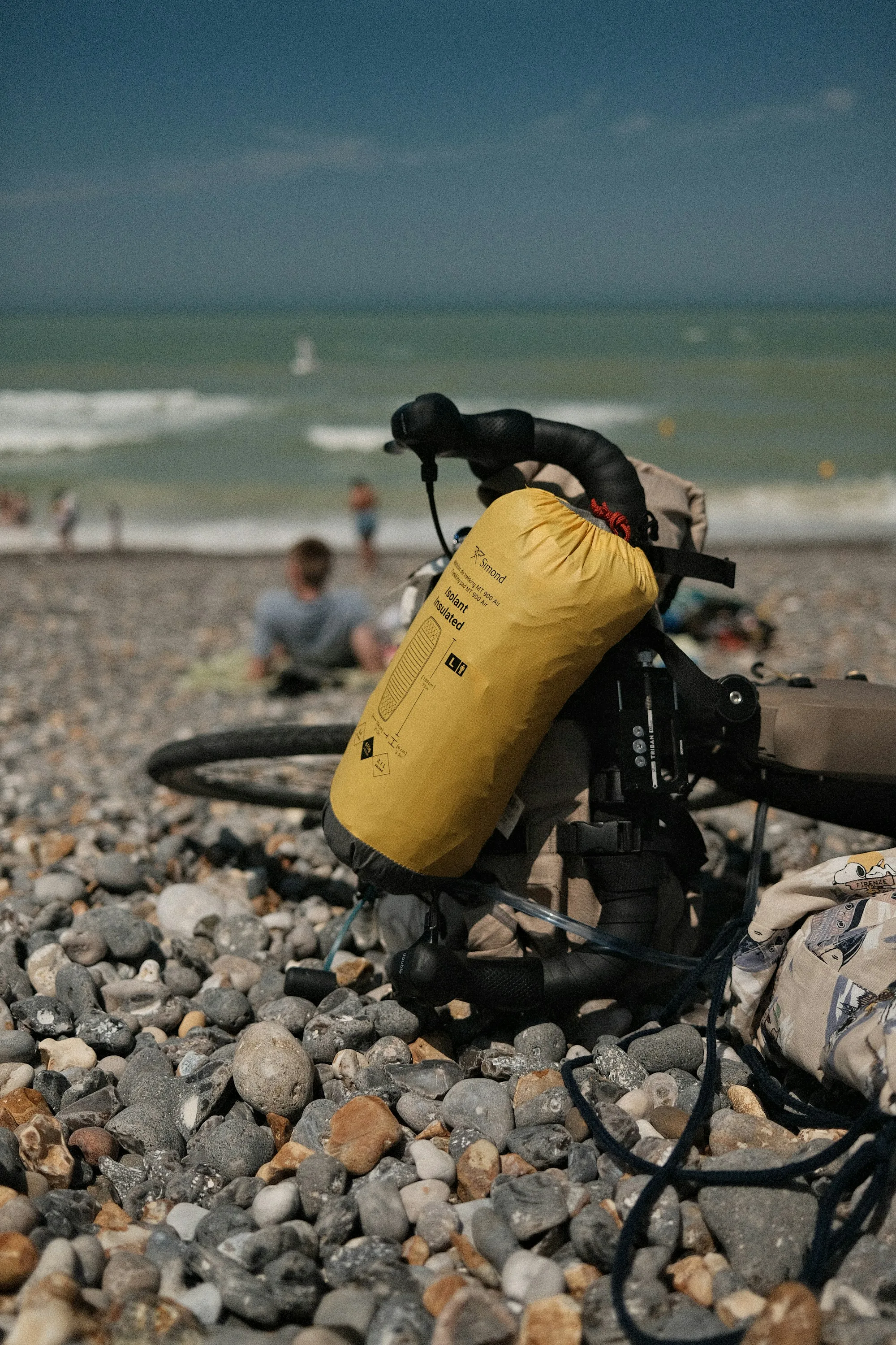 A yellow dry bag strapped to a loaded bike on a pebble beach