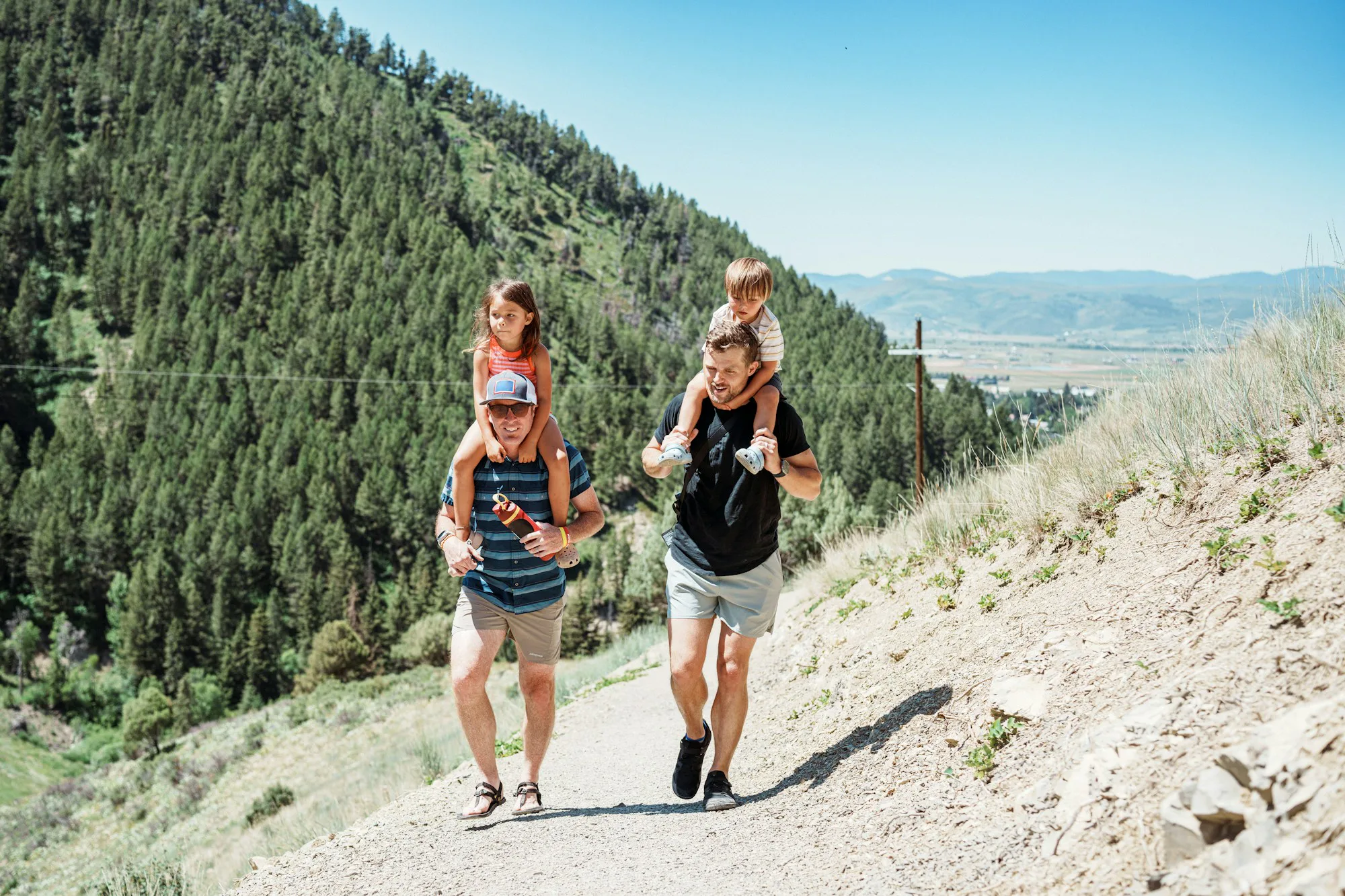 Two fathers carrying children on their shoulders on a mountain path
