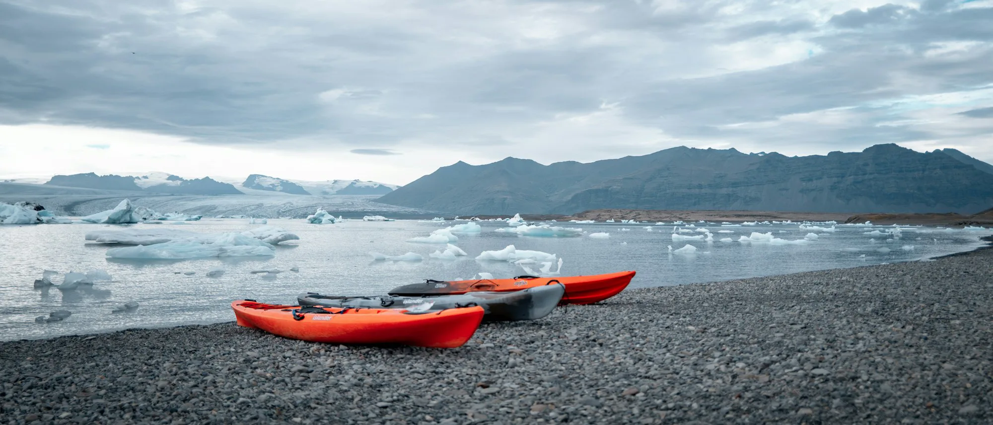Red kayaks on a dark shingle shore with icebergs beyond
