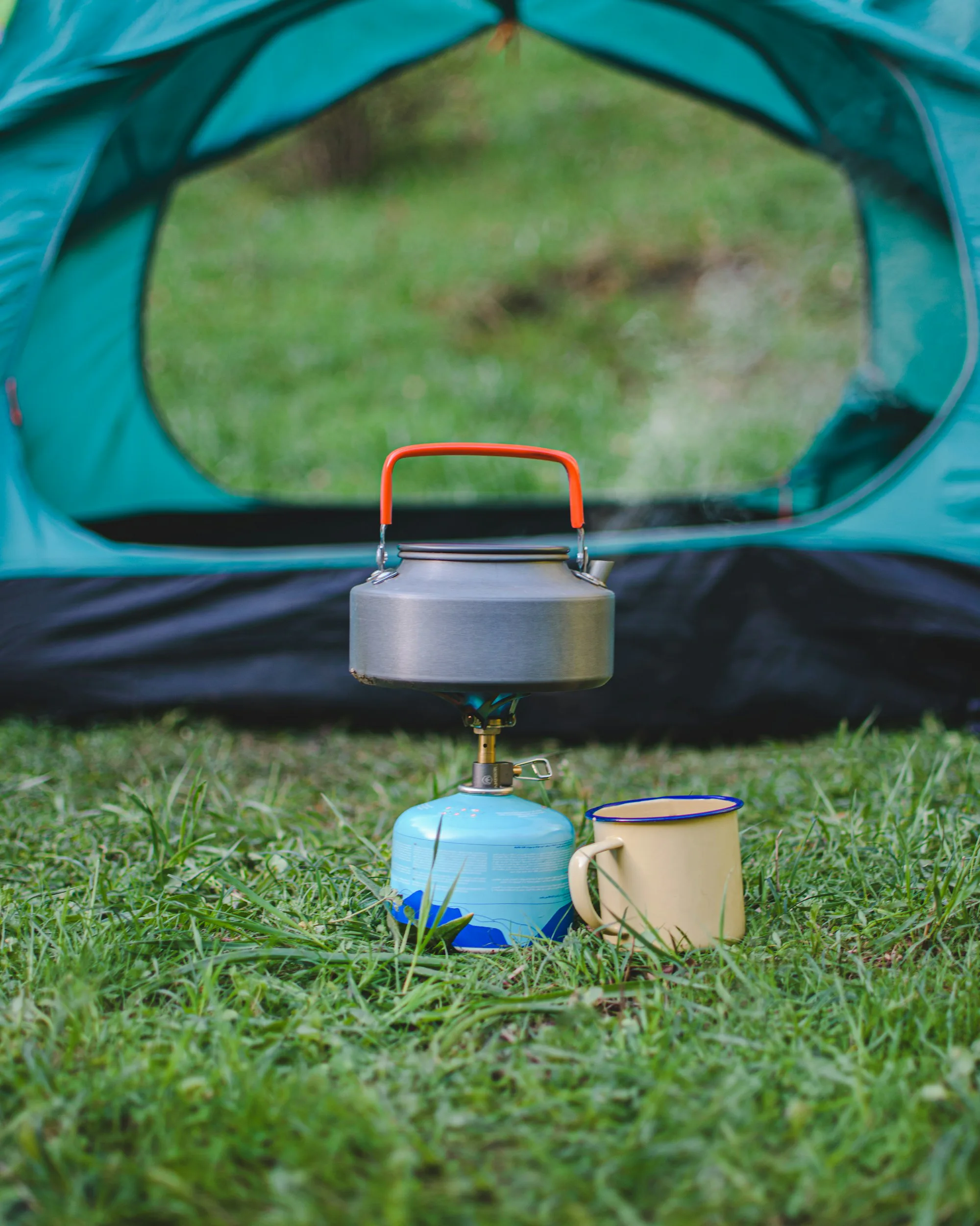 A kettle on a camping stove beside an enamel mug