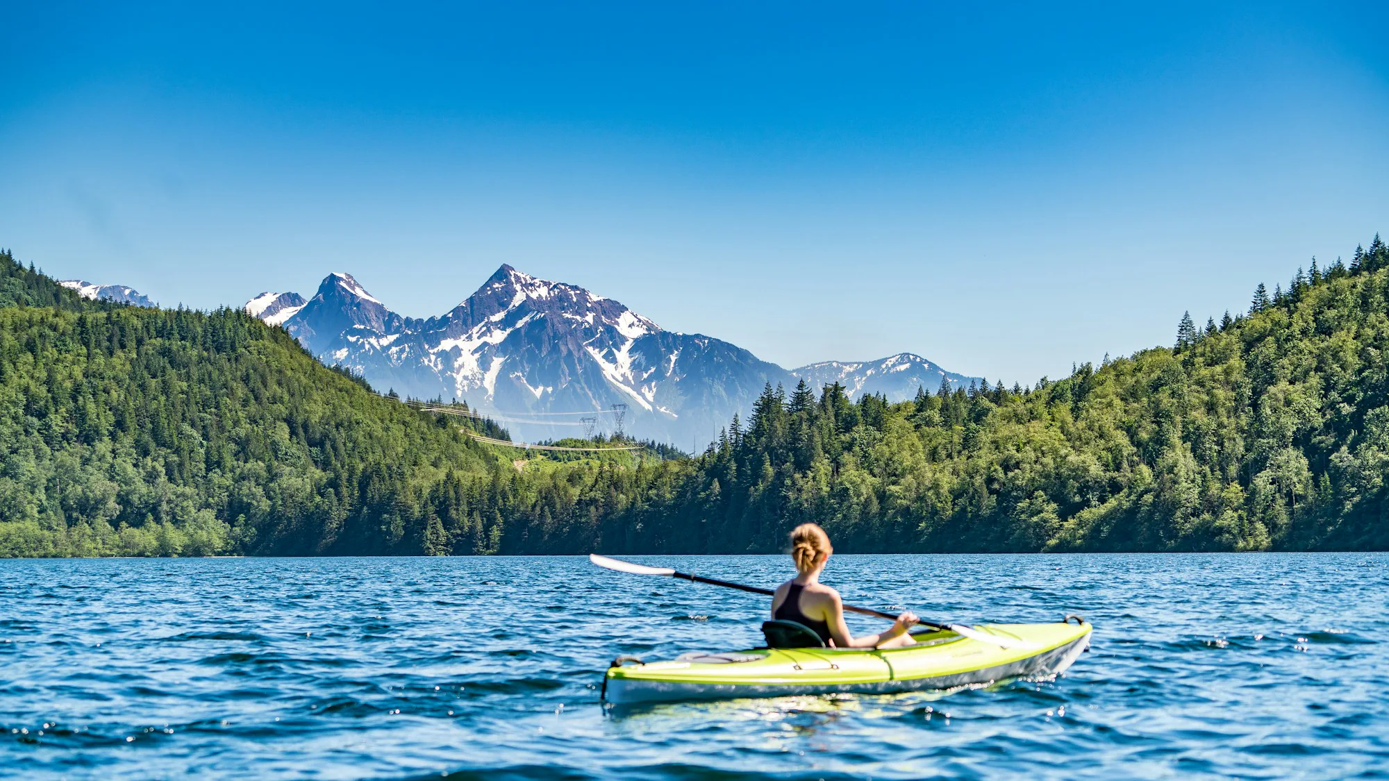 A paddler in a yellow kayak on a calm lake below forested hills and snowy peaks