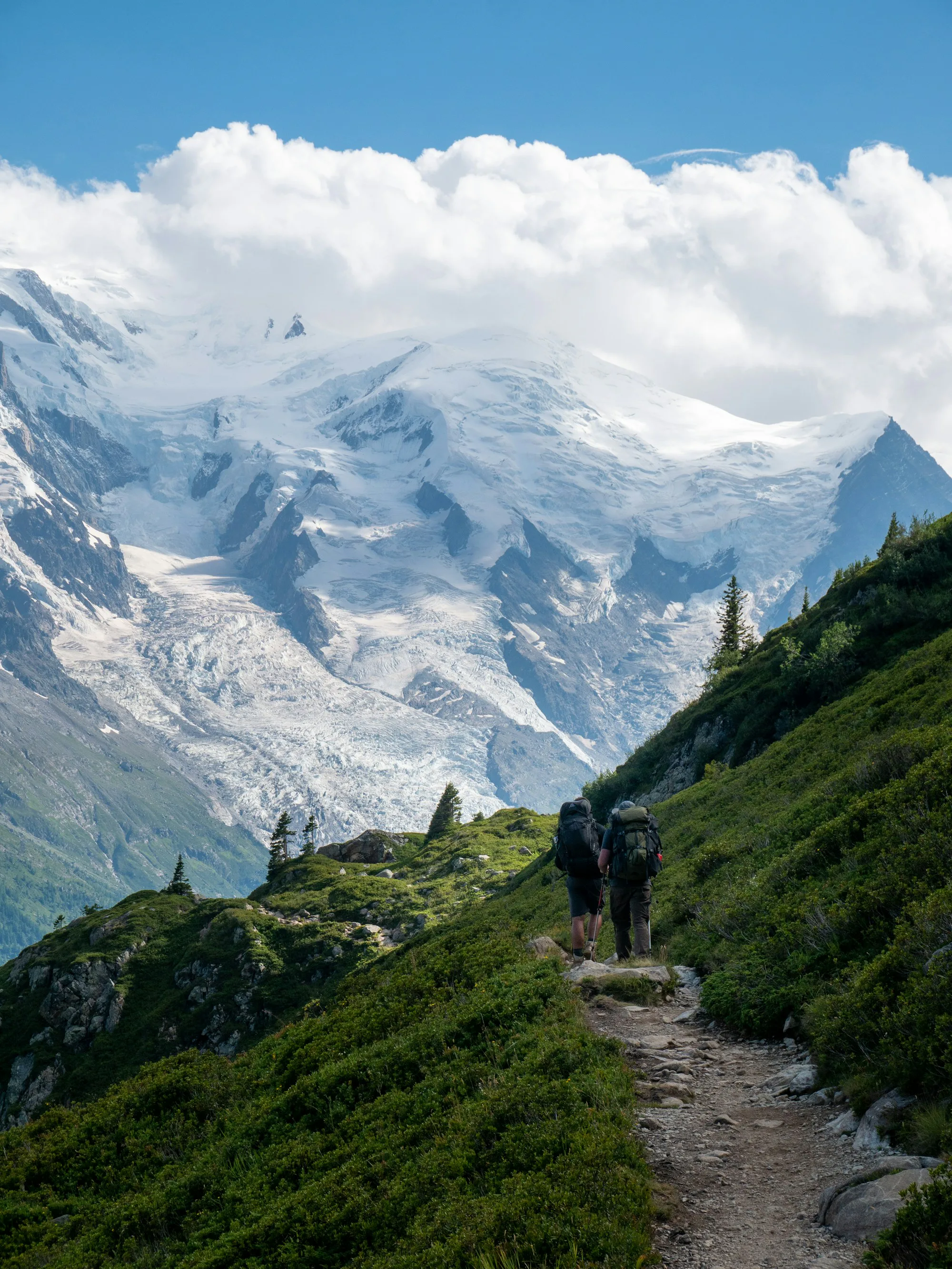 Hikers on a path above a glacier valley below snowy mountains