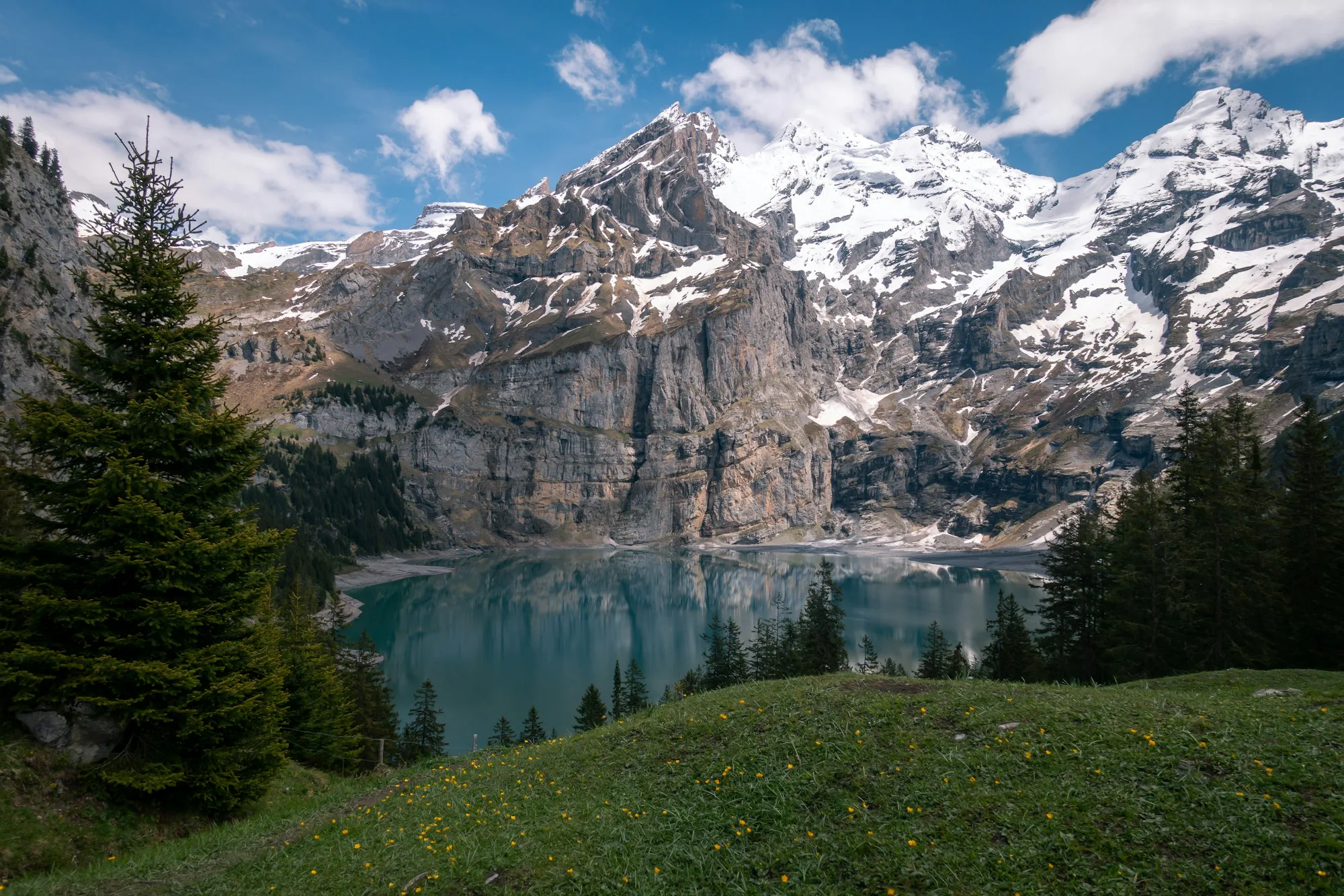 A turquoise mountain lake below rocky, snowy peaks and pine trees