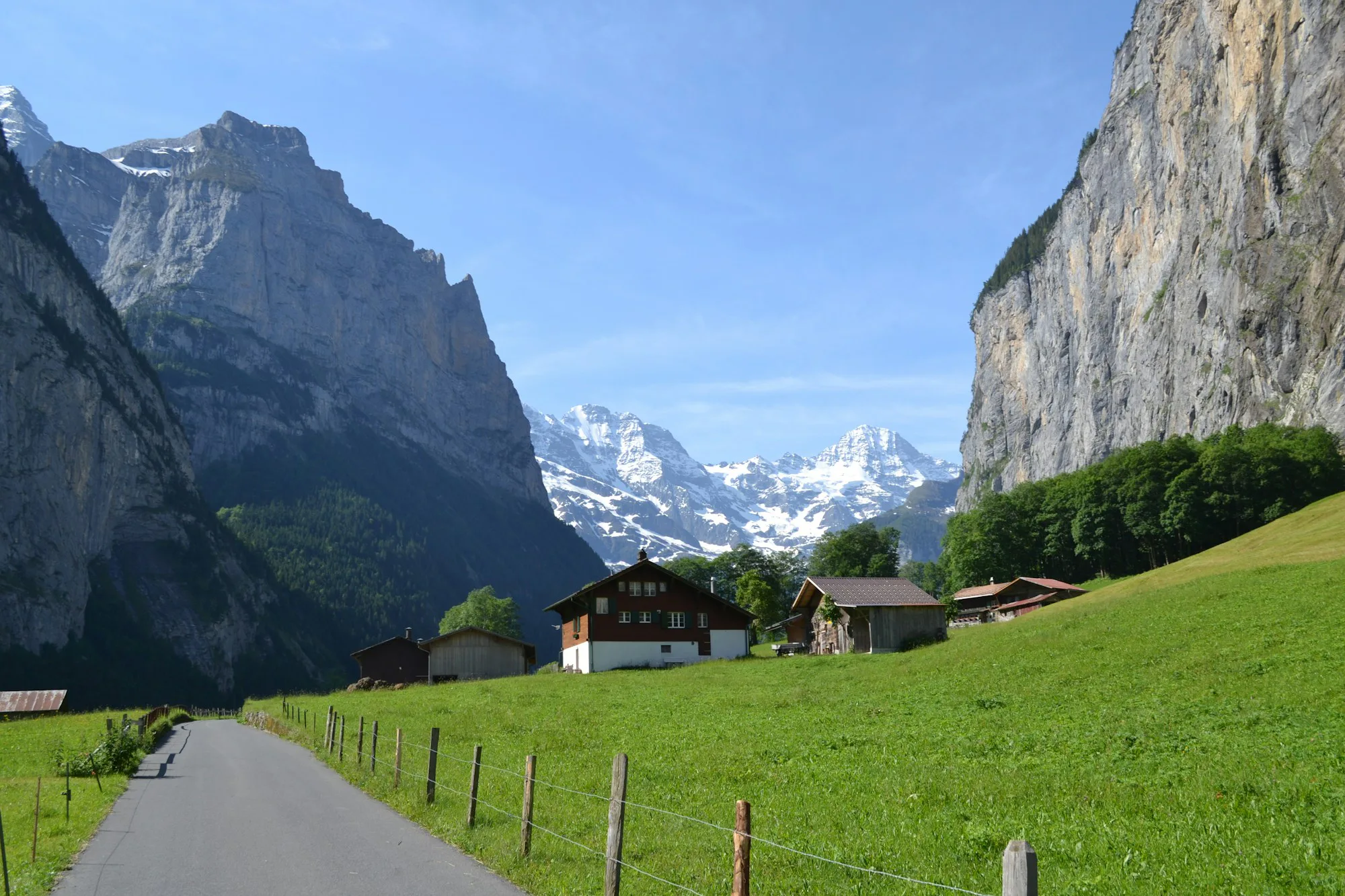 A lane through green meadows to chalets below a steep cliff